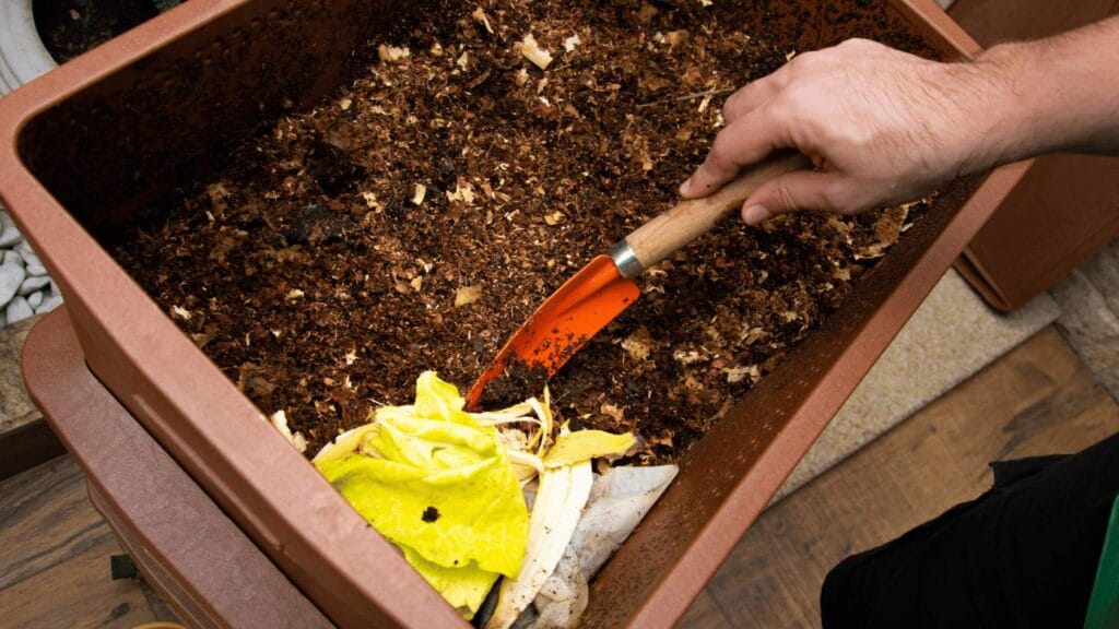 A person uses a small orange shovel to turn food scraps and soil in a compost bin. omahagardener.com