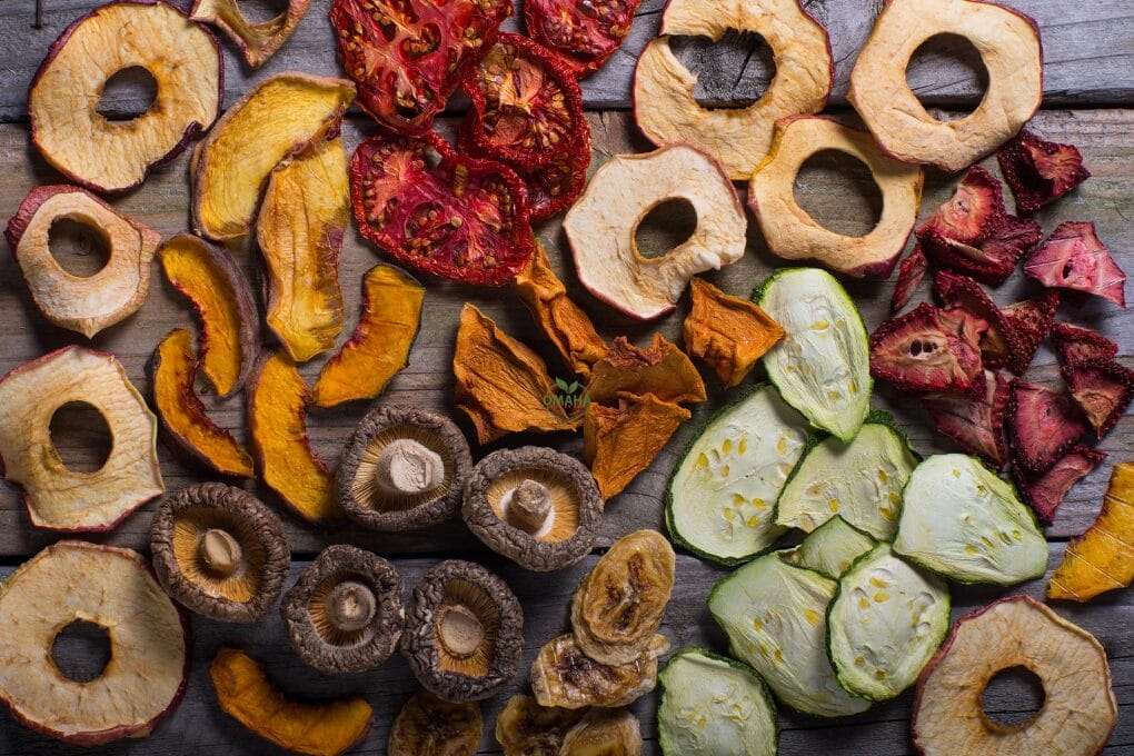 An assortment of dried fruits and vegetables including apple rings, tomato slices, peach slices, mushrooms, zucchini slices, and strawberries on a wooden surface made perfectly with a food dehydrator. omahagardener.com