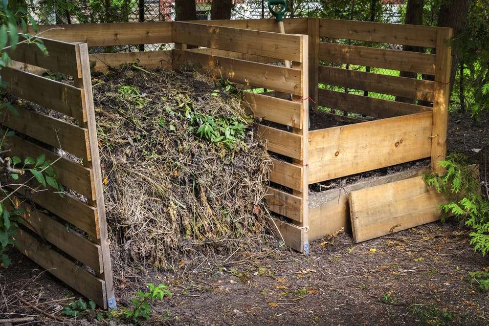 Can You Compost Pasta | Debunking Myths about Pasta and Rice 3 A wooden compost bin filled with garden waste, including dry leaves and branches for proper decomposition, sits outdoors in a shaded area surrounded by trees. A shovel is propped inside, ready for composting rice and other organic materials. omahagardener.com