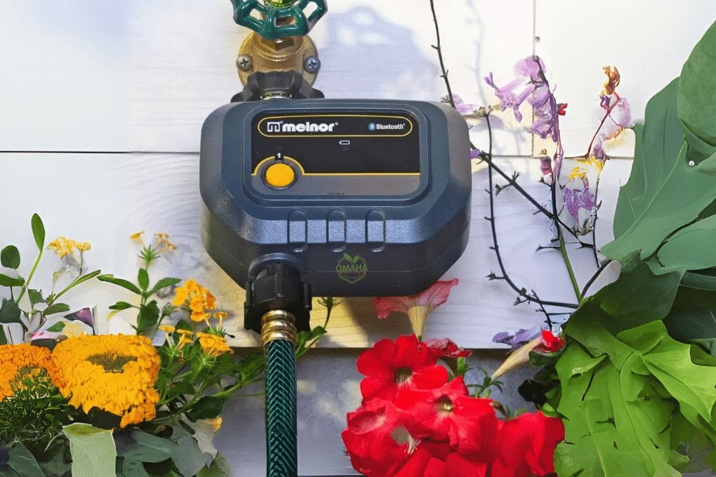 Melnor 65128-AMZ 4-Zone Bluetooth Water Timer with 5 Stainless Steel Filter Washers Set A Melnor Bluetooth water timer is attached to an outdoor faucet, surrounded by blooming flowers and green foliage. omahagardener.com