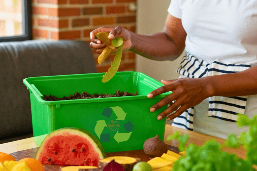 A person practices urban composting by placing fruit peels into a green recycling bin filled with compost, surrounded by assorted fruits and vegetables on a table. omahagardener.com